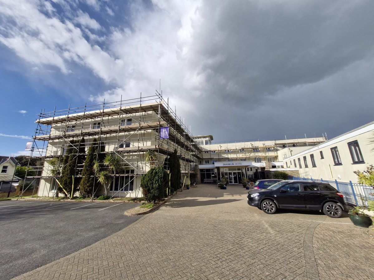 Modern building with extensive scaffolding under renovation, ivy-covered walls, parked car in foreground, partly cloudy sky